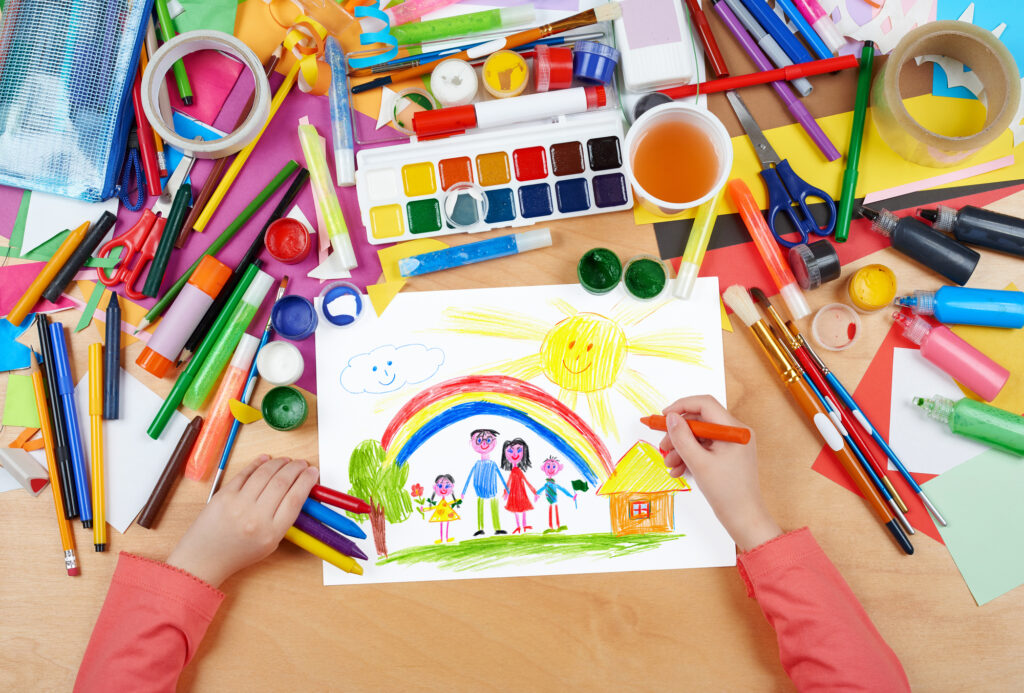 family on meadow with rainbow and house child drawing, top view hands with pencil painting picture on paper, artwork workplace